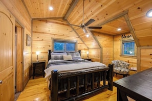 Une chambre dans une cabane en bois, avec un lit, une chaise, une table, un ventilateur de plafond et une fenêtre.