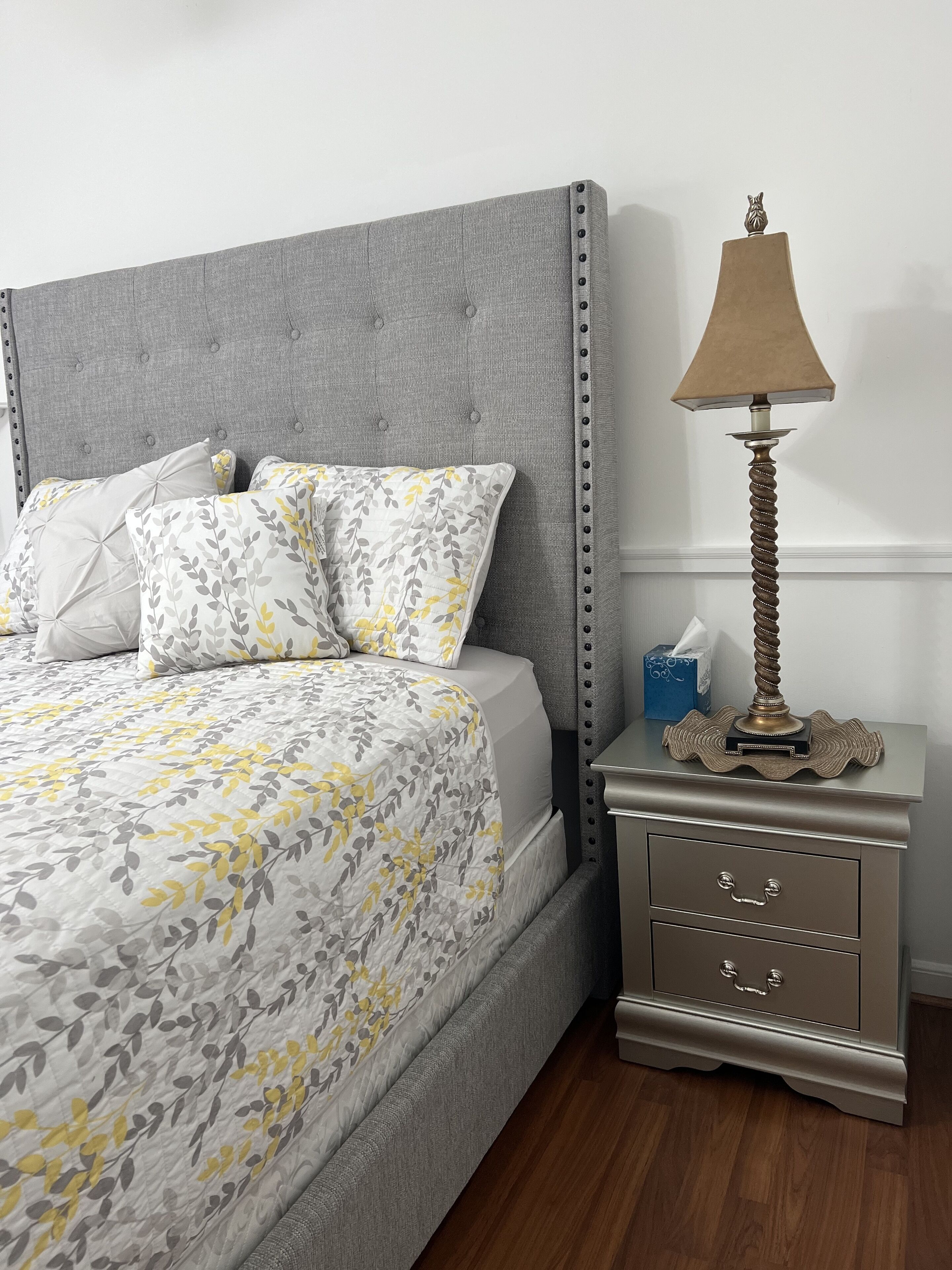 Une chambre à coucher avec une tête de lit rembourrée de couleur grise, un lit avec une literie à motifs floraux, une table de chevet avec une lampe et un petit objet décoratif.