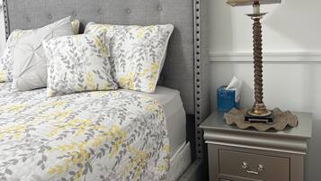 Une chambre à coucher avec une tête de lit rembourrée de couleur grise, un lit avec une literie à motifs floraux, une table de chevet avec une lampe et un petit objet décoratif.