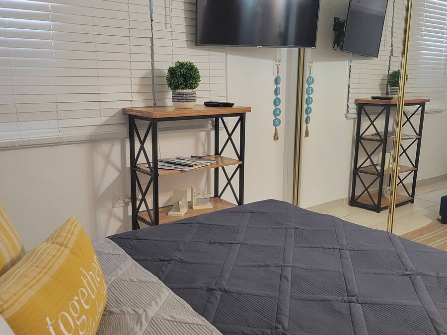 Un dormitorio con una cama, un televisor montado en la pared y dos mesitas con adornos.