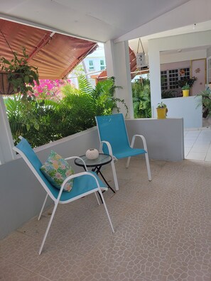 A covered outdoor seating area with two blue chairs and a small table, surrounded by potted plants and a view of a building. - Playa icacos (Fajardo)