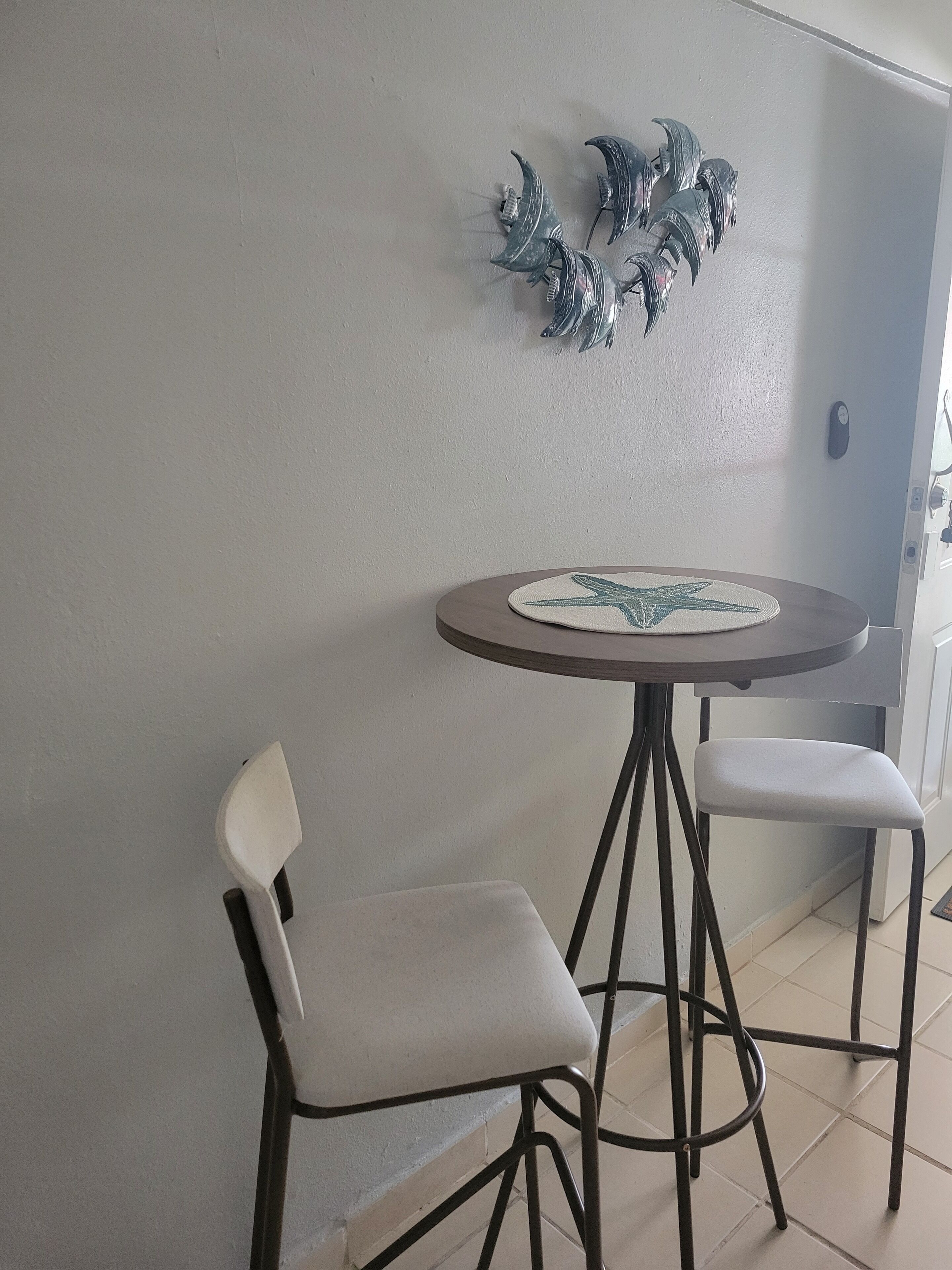 A small round table with a placemat, two white chairs with metal frames, and a wall-mounted decorative piece.