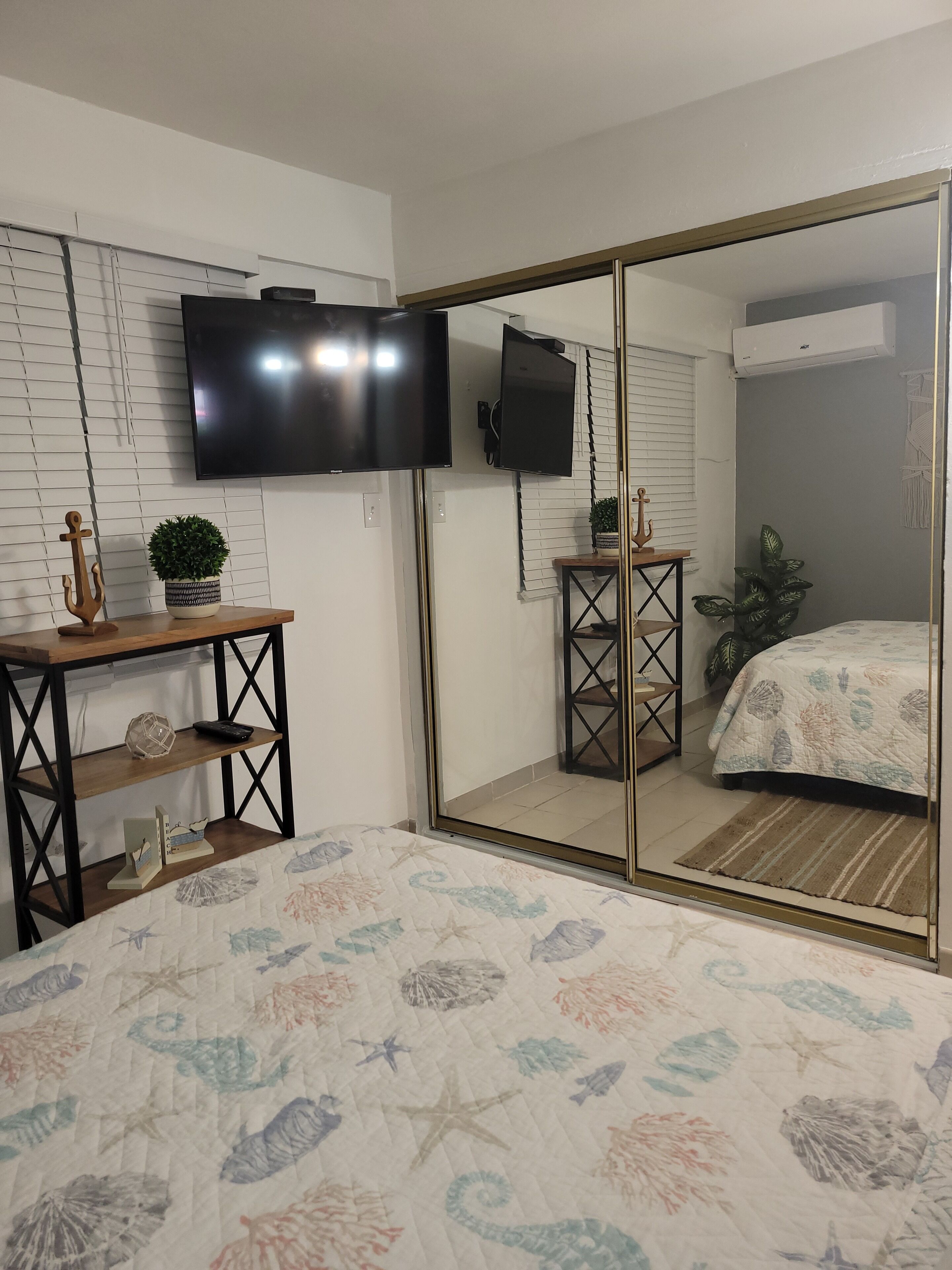 A bedroom with a bed, a TV mounted on the wall, a small table with a plant, and a mirror.