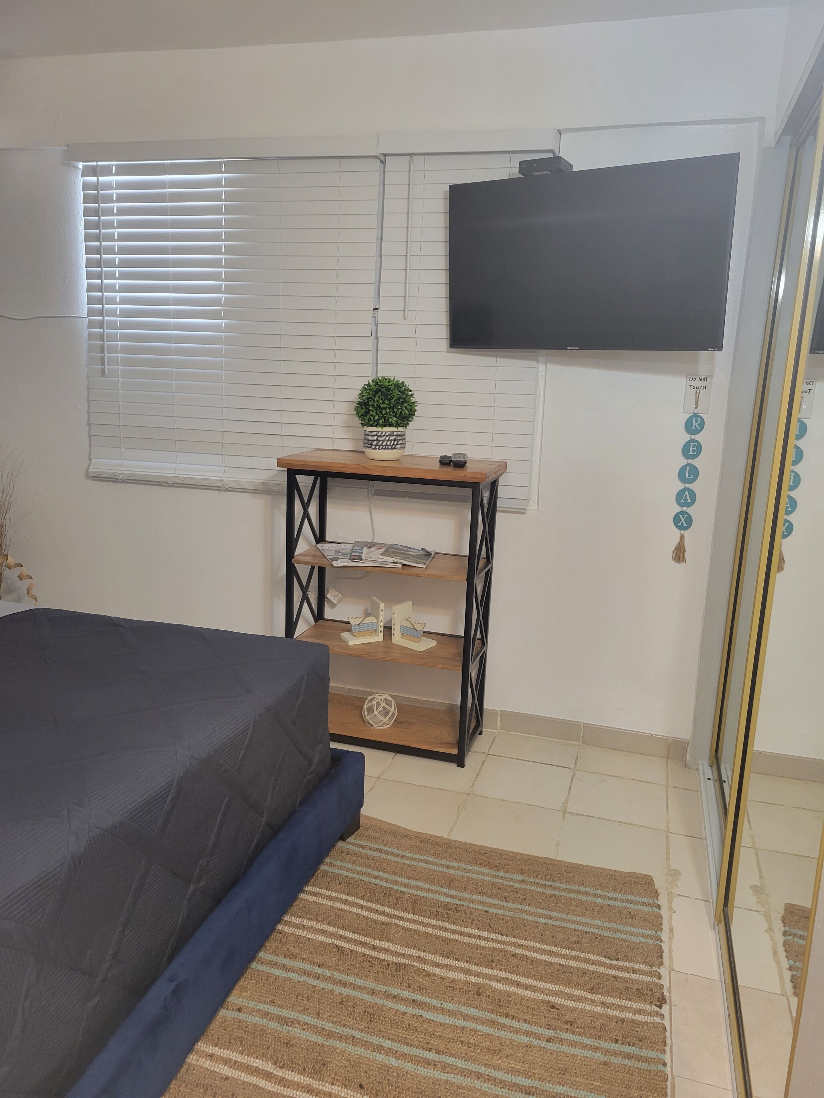 Un dormitorio con una cama, un televisor montado en la pared, una mesita con una planta y un estante con adornos.