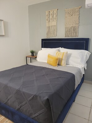 A neatly made bed with a dark grey quilt, white and yellow pillows, and a blue headboard. There are two macramé wall hangings above the bed. - Playa icacos (Fajardo)