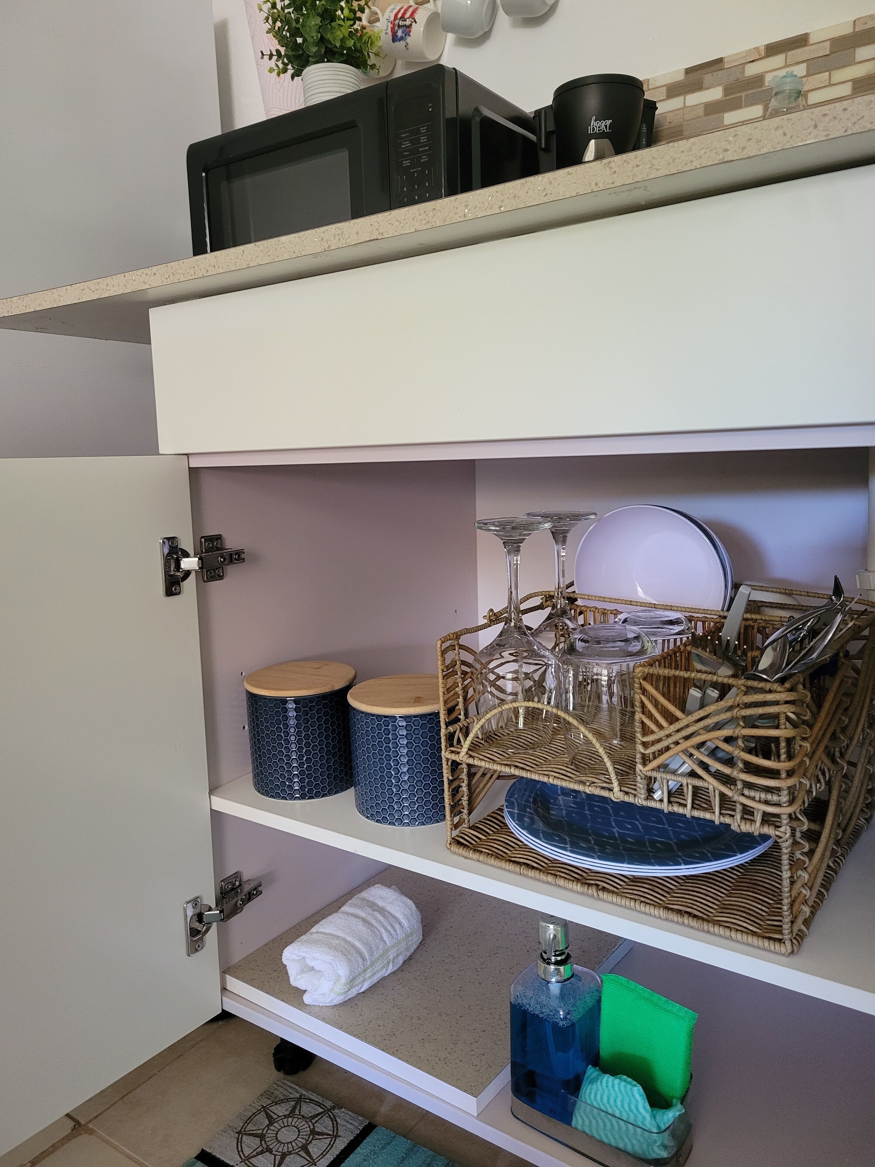 A kitchen with a microwave, two blue patterned containers, a wicker basket with glassware, a white towel, and a blue bottle.