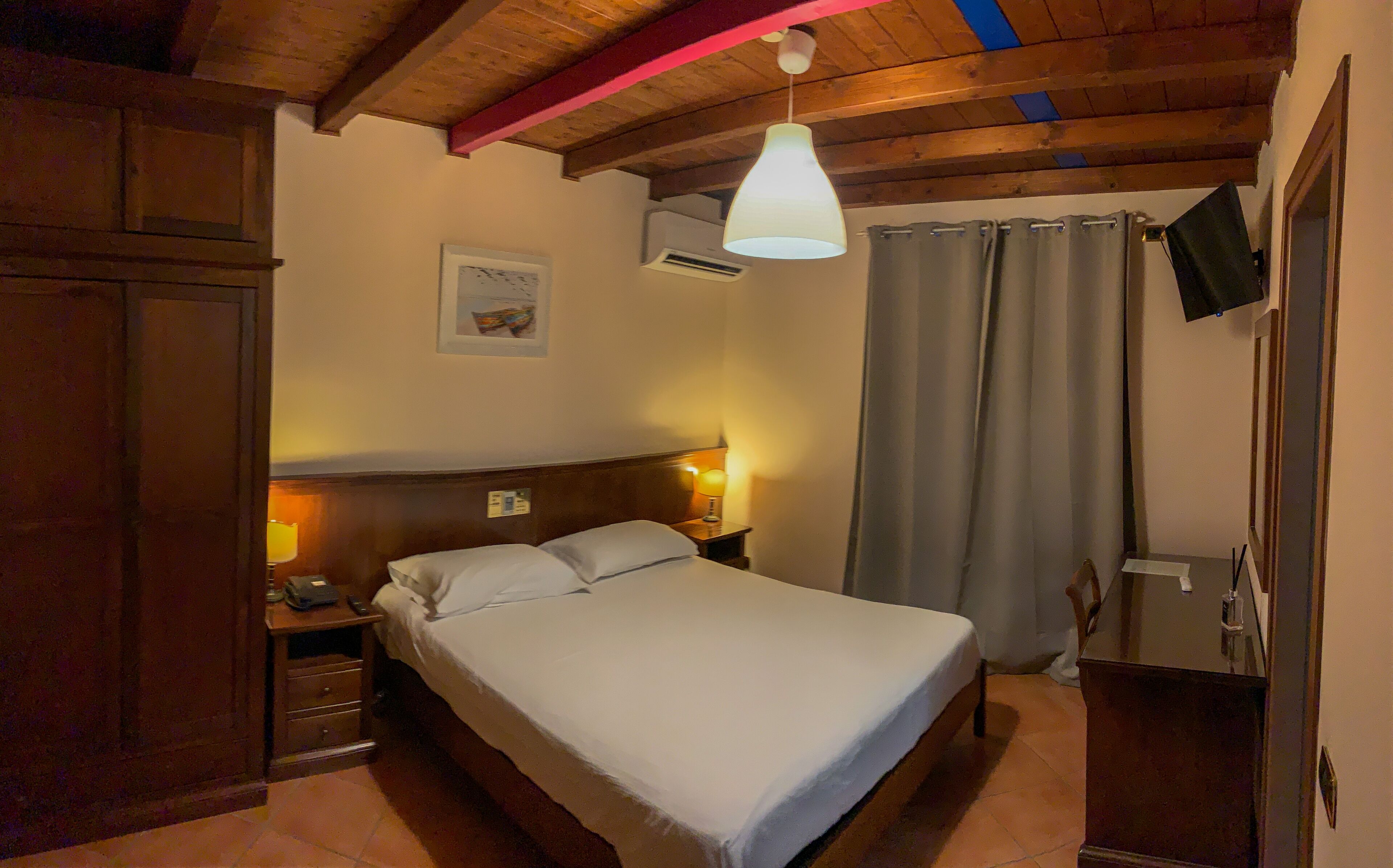 A bedroom with a bed, bedside tables, a nightstand, a wall-mounted air conditioner, a ceiling light fixture, a framed picture, and a door with a towel hanging on it.