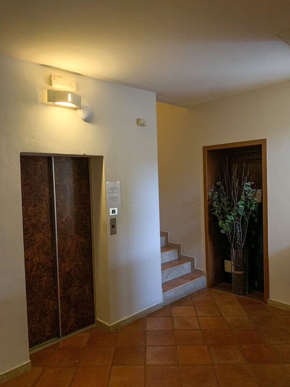 Un pasillo interior con un ascensor de mármol, un aplique de pared, una escalera y una planta en maceta.