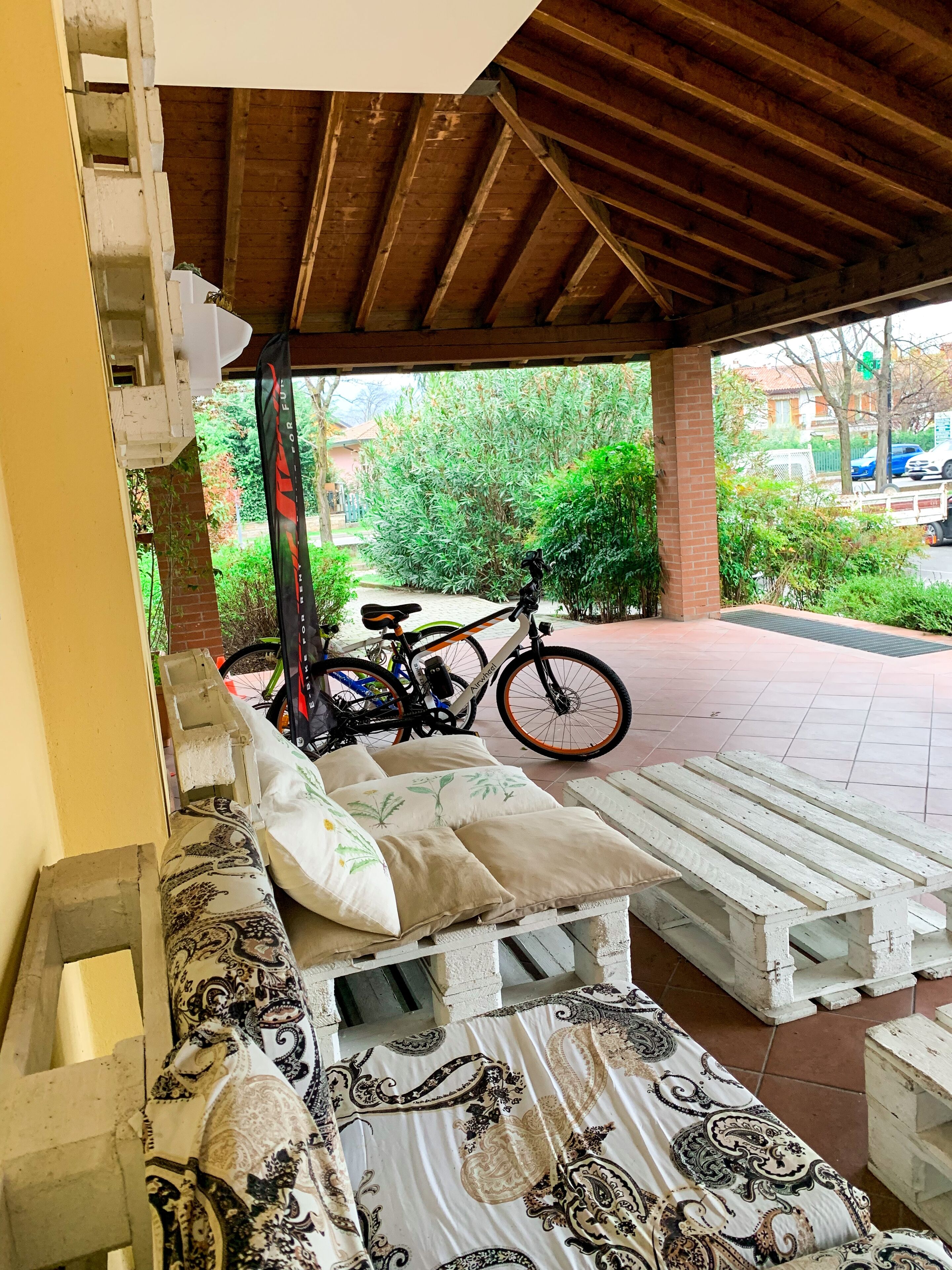 Een overdekt terras met een bank en kussens, een fiets, en een uitzicht op groen en gebouwen op de achtergrond.