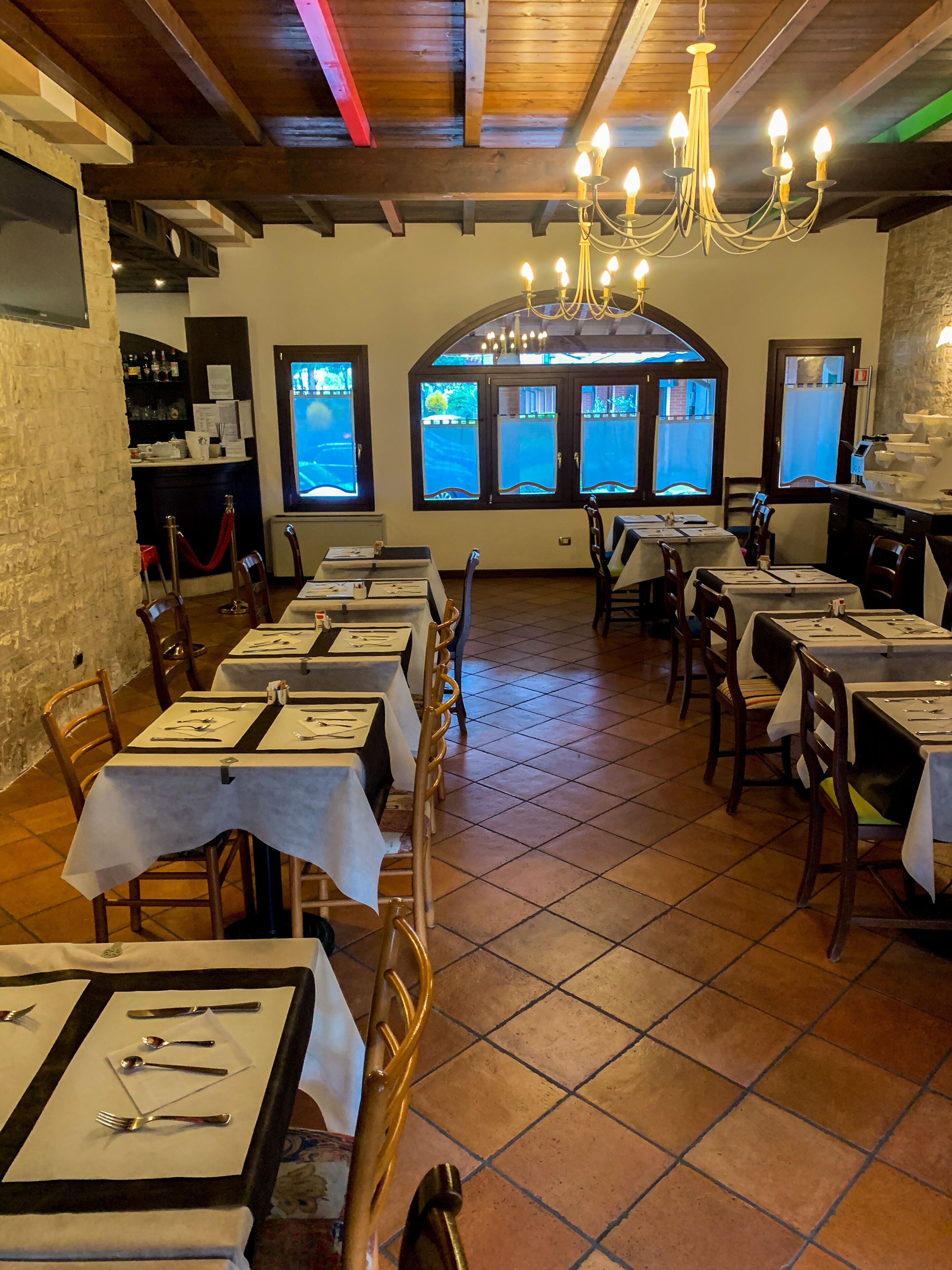 Un restaurante con varias mesas, sillas y un candelabro. Hay ventanas con vistas al exterior.