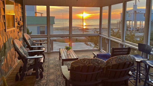 A sunlit room with large windows overlooking a beach at sunset, wooden furniture, and a ceiling fan. - 5 Bedroom Beach House Slps 12 - Private Beach - Shells - Fishing -Screened porch (Panacea)