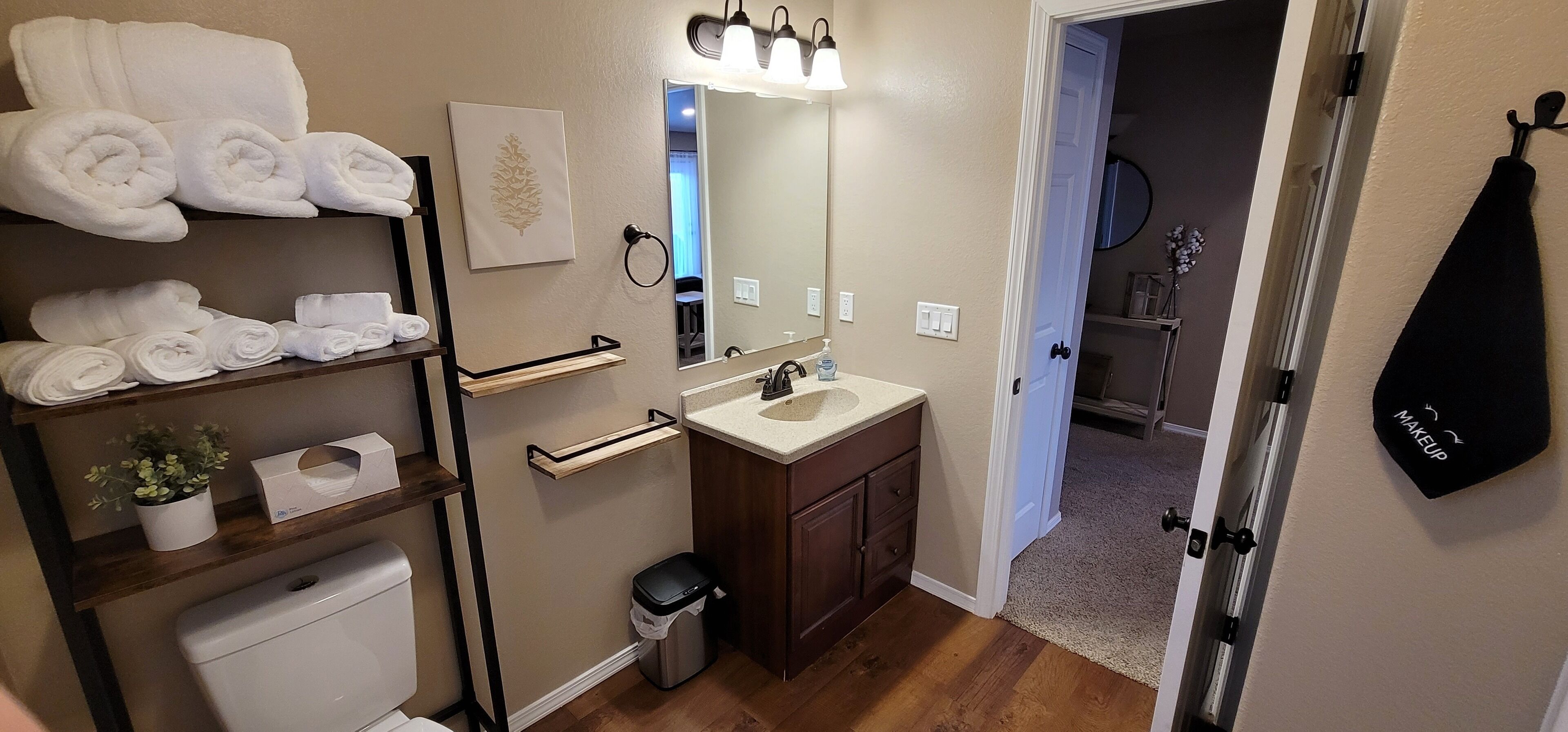 A bathroom with a toilet, sink, and towel rack. There is a mirror above the sink and a towel rack with folded towels. A trash can is visible next to the toilet.