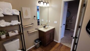 A bathroom with a toilet, sink, and towel rack. There is a mirror above the sink and a towel rack with folded towels. A trash can is visible next to the toilet.