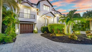 A two-story house with a front entrance, a covered porch, and a driveway leading to a garage. - Tern Key - Luxury Canal-Front Home w/ Heated Pool, Spa, Dock & Near Bean Point (Anna Maria)