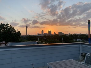 Terraço no telhado com horizonte urbano ao pôr do sol, incluindo uma torre imponente e edifícios altos.