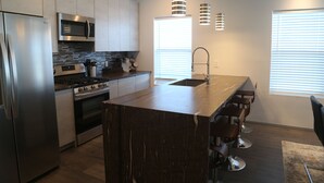 A modern kitchen with stainless steel appliances, a central island with bar stools, pendant lighting, and a large window providing natural light. - Modern Home With Rooftop Patio - Down Town Views (San Antonio)