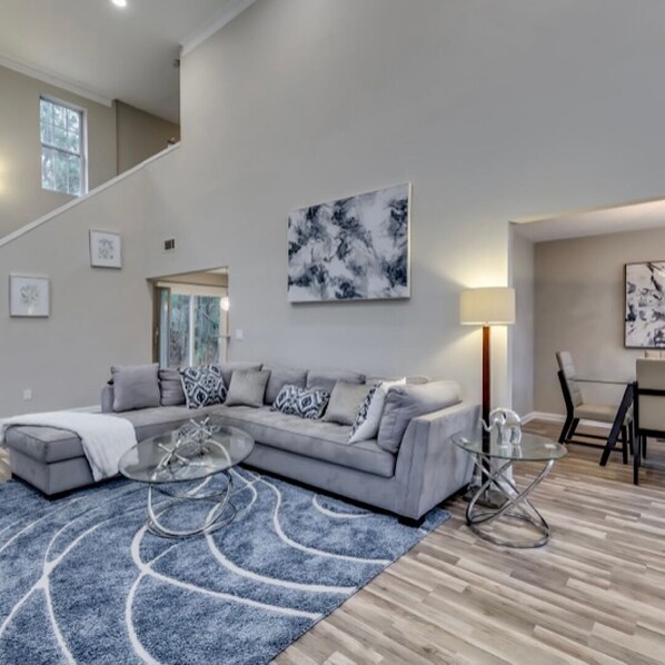 A modern living room with a grey sectional sofa, a glass coffee table, and a blue patterned rug.