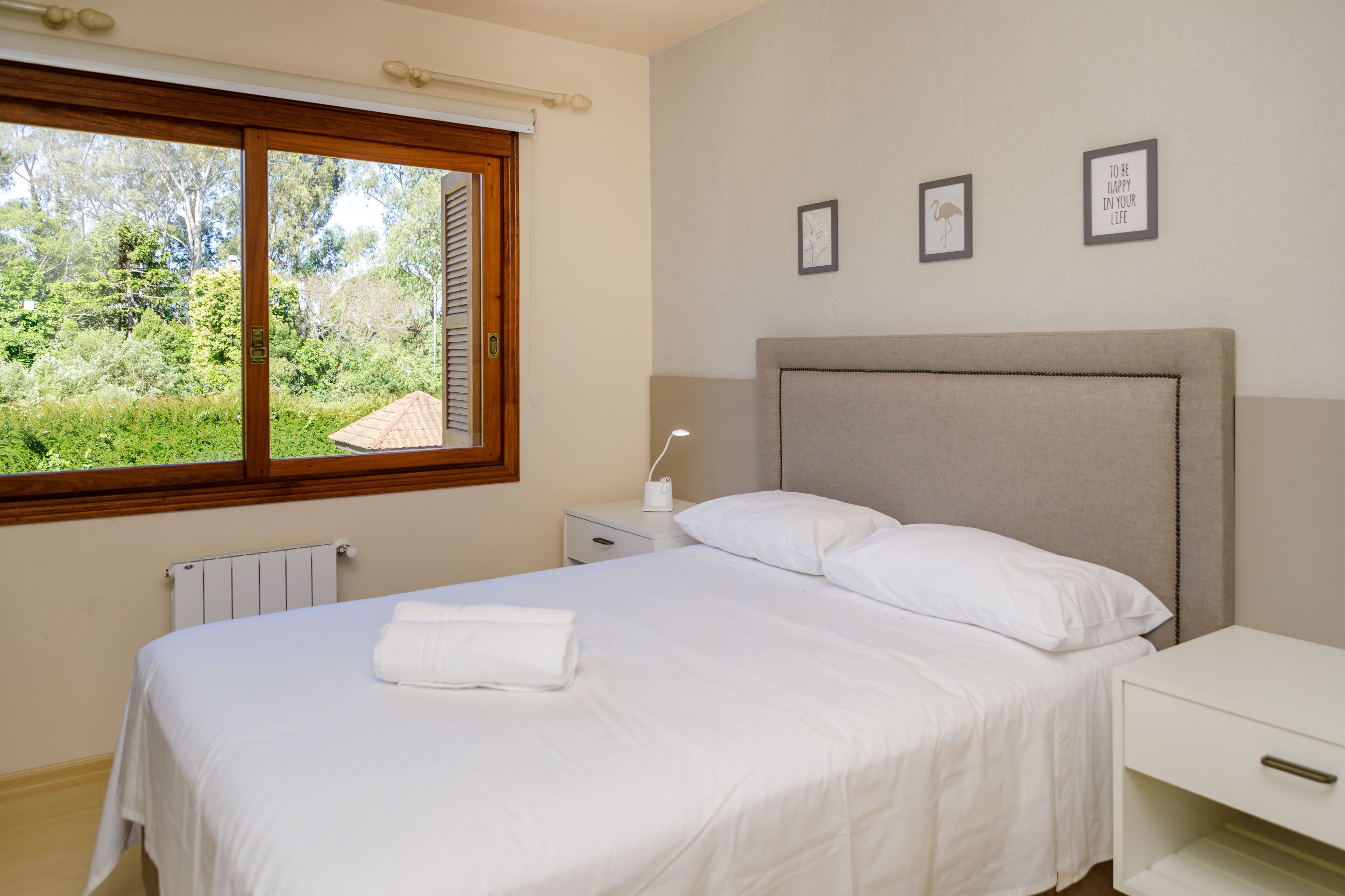 A bedroom with a bed, a bedside table, a nightstand, a window with blinds, and a radiator.