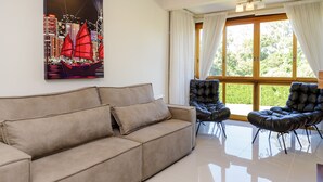 Un salon moderne avec un canapé beige, deux fauteuils en cuir noir, une grande fenêtre avec des rideaux et un tableau représentant un paysage urbain accroché au mur.