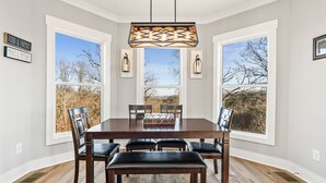 Une salle à manger avec une table en bois, six chaises et un lustre. Deux fenêtres offrent une vue sur les arbres et un mur en pierre.