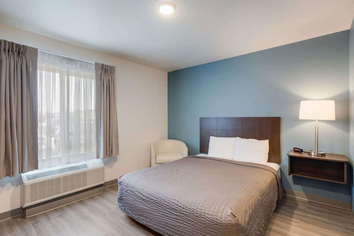 Een hotelkamer met een bed, een stoel, een nachtkastje met een lamp en een raam met gordijnen.