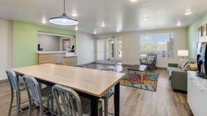 A modern kitchen with a wooden table and chairs, a dining area with a patterned rug, and a living space with a sofa and lamp.