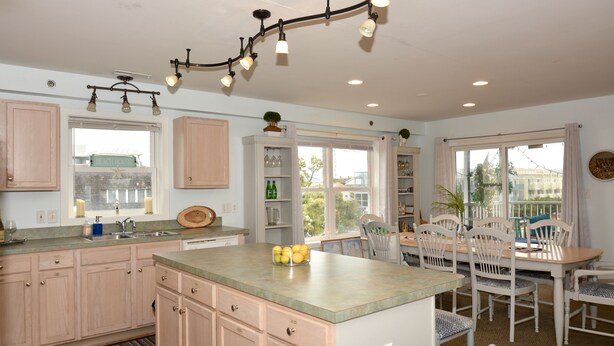 A kitchen with a central island, wooden cabinets, a dining area with white chairs, and a window with a view of the outside.
