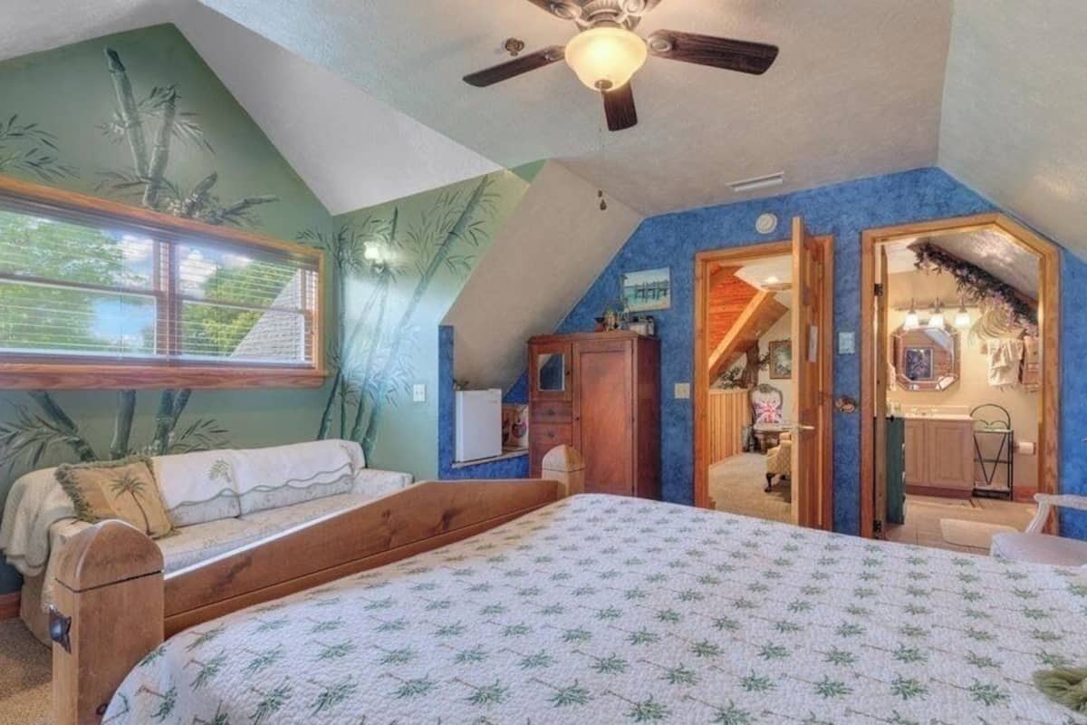 Een slaapkamer met een groot bed, een raam met zonwering, een plafondventilator en een deur naar een andere kamer.