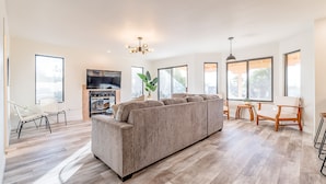 Ein geräumiges Wohnzimmer mit einer großen grauen Couch, Holzböden und großen Fenstern, die für natürliches Licht sorgen.
