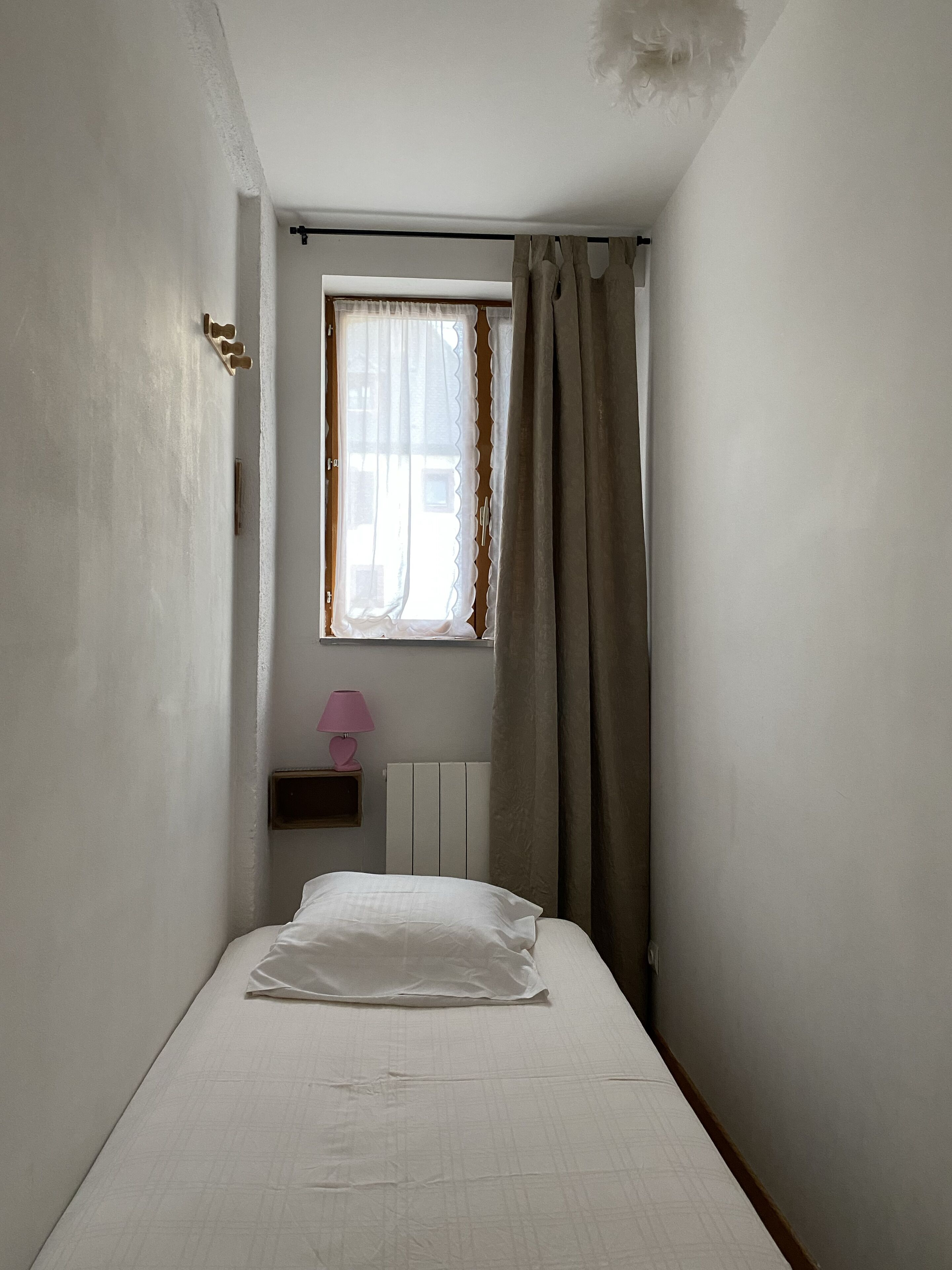 A small, narrow room with a single bed, a window with curtains, a radiator, and a pink lamp on a shelf.