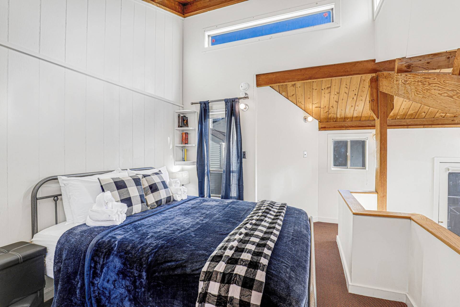 Un dormitorio con cama, una mesita de noche, estante con libros, ventana y estructura de techo de madera.