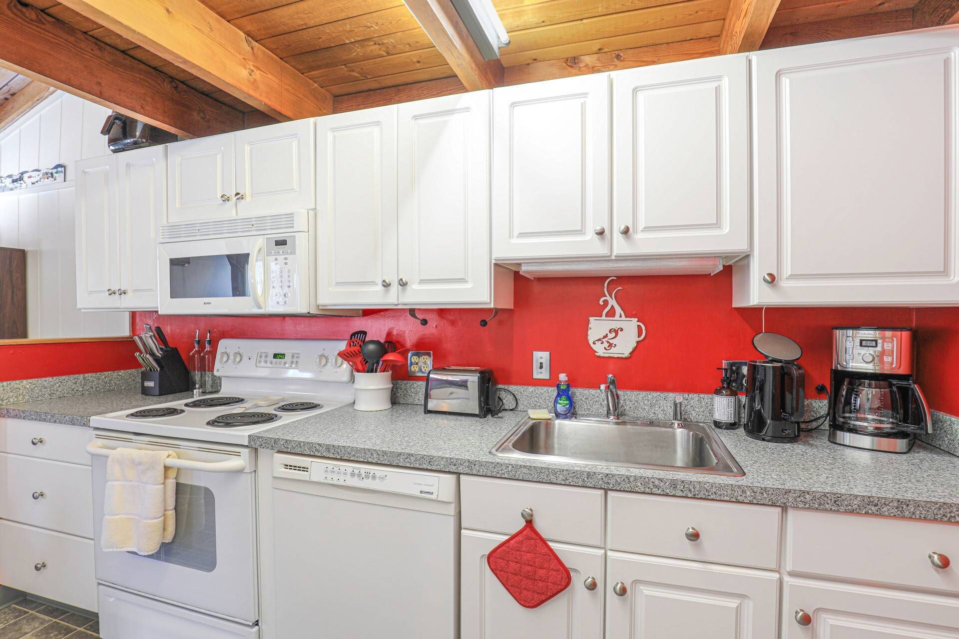 A kitchen with white cabinets, a red backsplash, a stove, a microwave, a dishwasher, and a coffee maker.