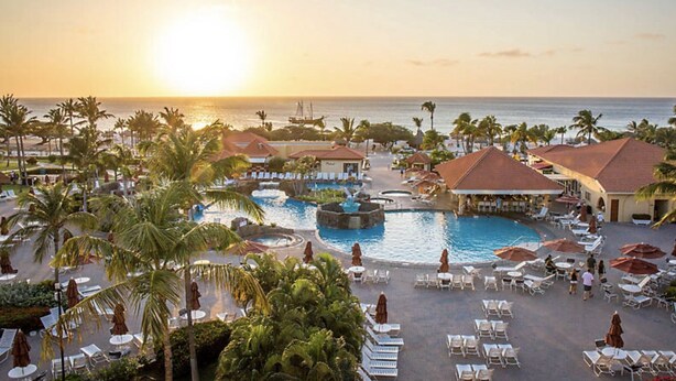 Ein Resort mit einem zentralen Pool, Liegebereichen und Blick auf den Sonnenuntergang über dem Meer.