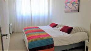 Ein Schlafzimmer mit einem Bett, einer bunten Decke, zwei roten Kissen, einer weißen Wand und einem Gemälde über dem Bett.