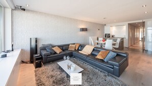 A modern living room with a sectional sofa, a coffee table, and a TV. There is a kitchen in the background. - Marble Floor, full comfort sea view, parking incl. (Oostende)