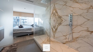 A modern bathroom with a marble wall, a glass shower enclosure, and a visible bed in the adjacent room. - Marble Floor, full comfort sea view, parking incl. (Oostende)