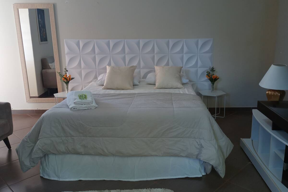 Una cama bien tendida con cabecera blanca, una mesita de noche con una lámpara y un mueble bajo para televisión con control remoto.