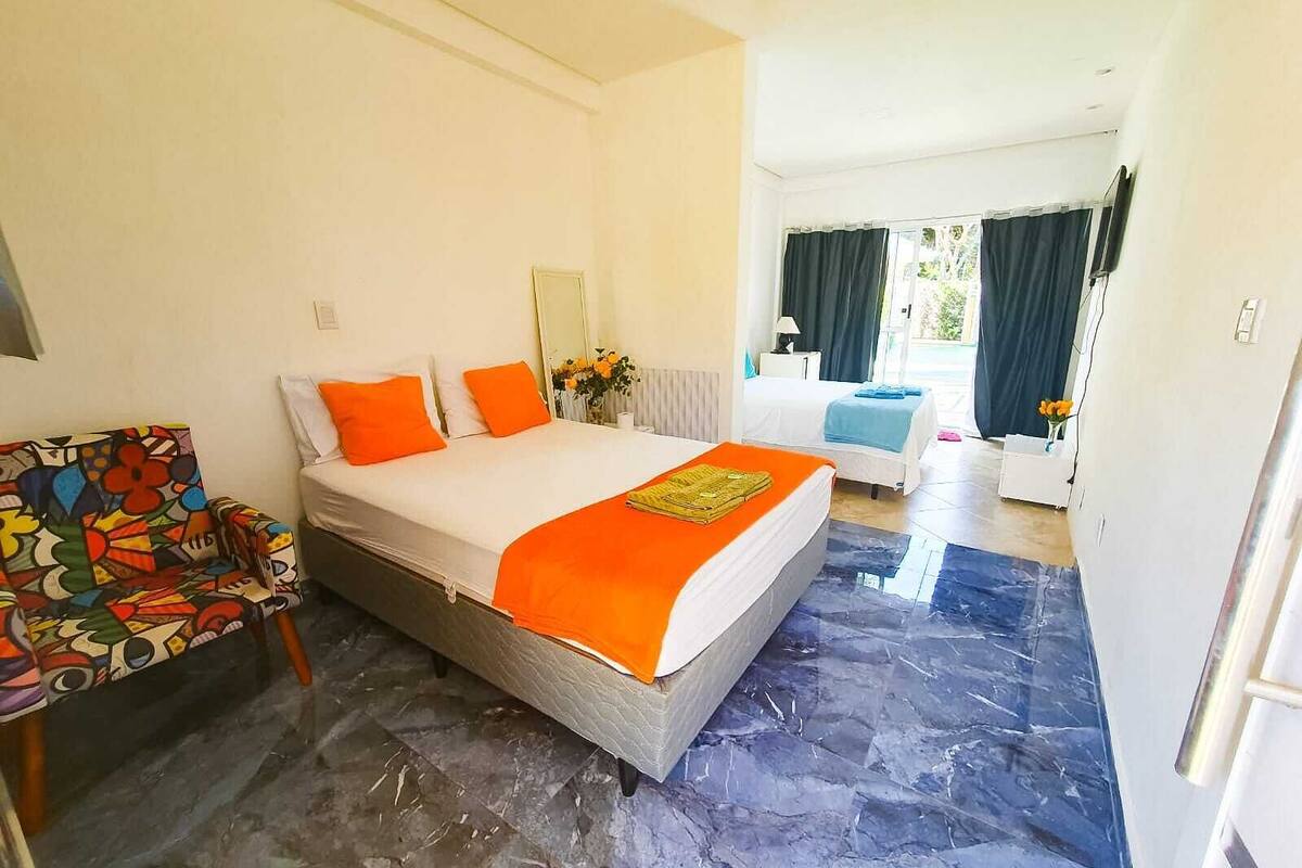 Una habitación de hotel con cama, una silla colorida, una mesita y un baño visible a través de una puerta abierta.