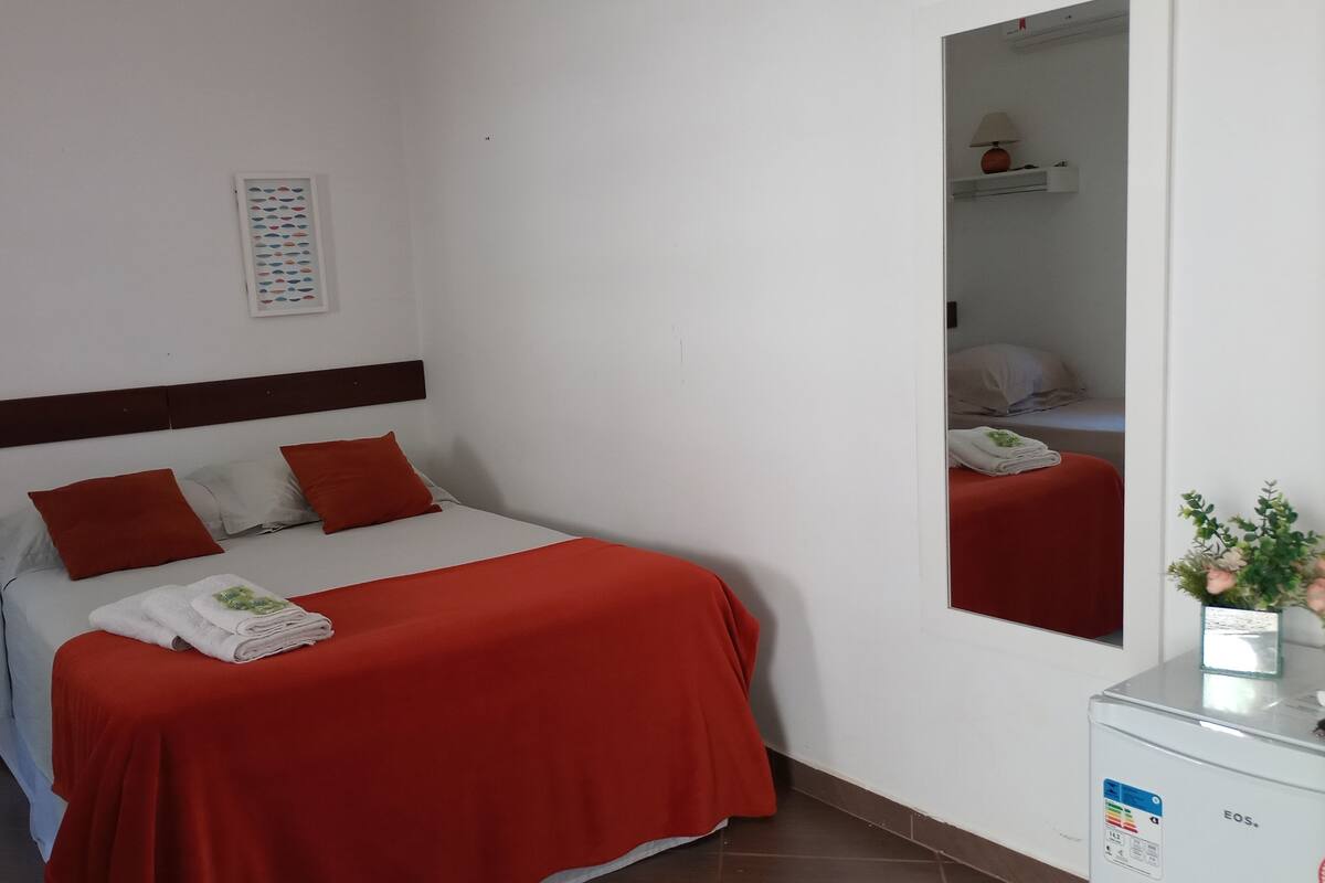 Habitación de hotel con cama, ropa de cama roja, cabecero blanco, un espejo y un mueble pequeño con una planta.