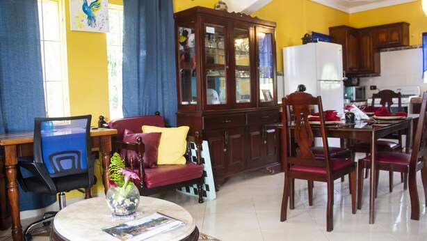 A dining area with a wooden table, chairs, and a red sofa. There is a cabinet with glass doors and a white refrigerator. - El Castilito (Brown's Town)