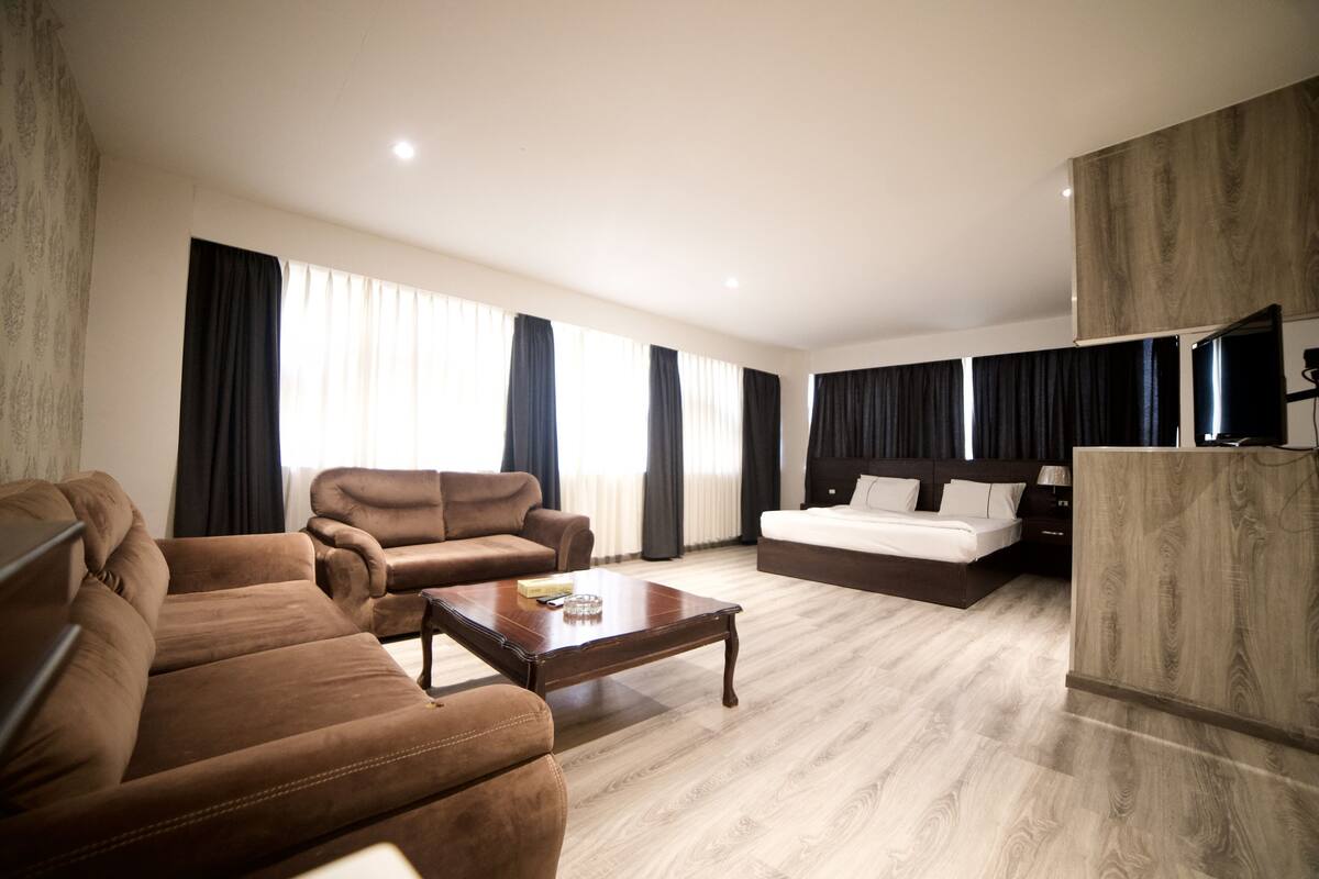 Une chambre d’hôtel moderne avec un canapé en cuir marron, une table basse en bois, un grand lit avec une tête de lit sombre et un téléviseur posé sur une commode en bois.