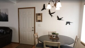 A dining area with a round table, chairs, and a chandelier. There are wall decorations of birds and a door with a sign.