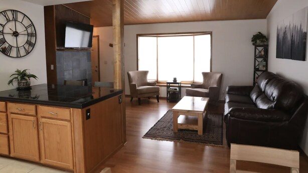 A modern kitchen with wooden cabinets and a black countertop, a dining area with a wooden table and chairs, and a living area with a leather couch and a large window.