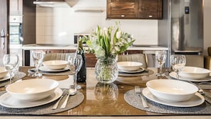 A modern kitchen with wooden cabinets, a stainless steel refrigerator, and a dining table set for six with white plates, glasses, and a vase of flowers.