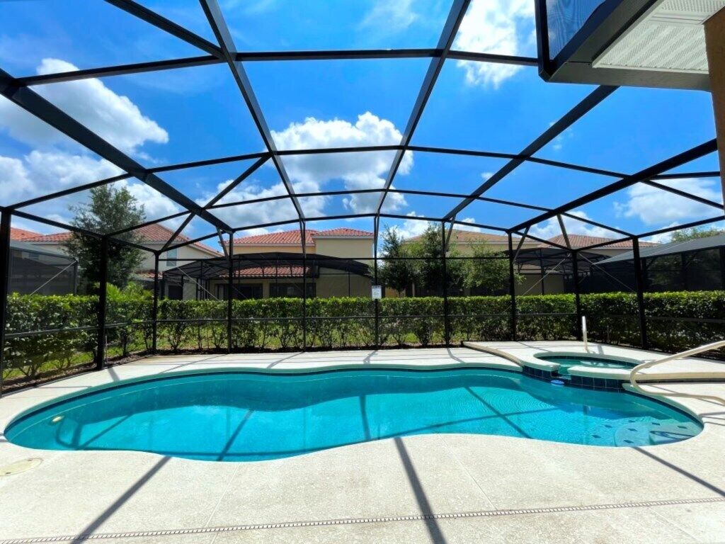 A swimming pool with a clear blue water, surrounded by a covered area with a metal frame and a view of houses in the background.