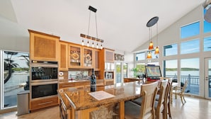A spacious kitchen with granite countertops, wooden cabinets, and a large island. There is a dining area with a table and chairs, and large windows offering a view of the outdoors.