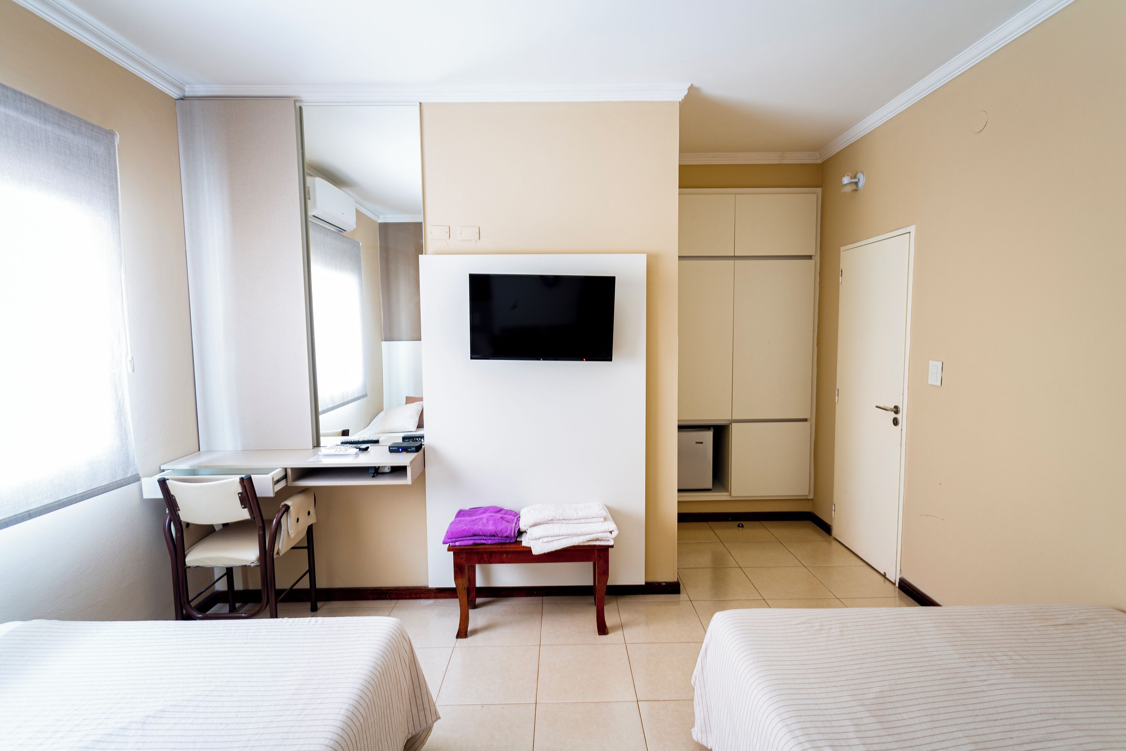 A hotel room with two beds, a desk with a chair, a TV mounted on the wall, and a bathroom with a toilet and sink.