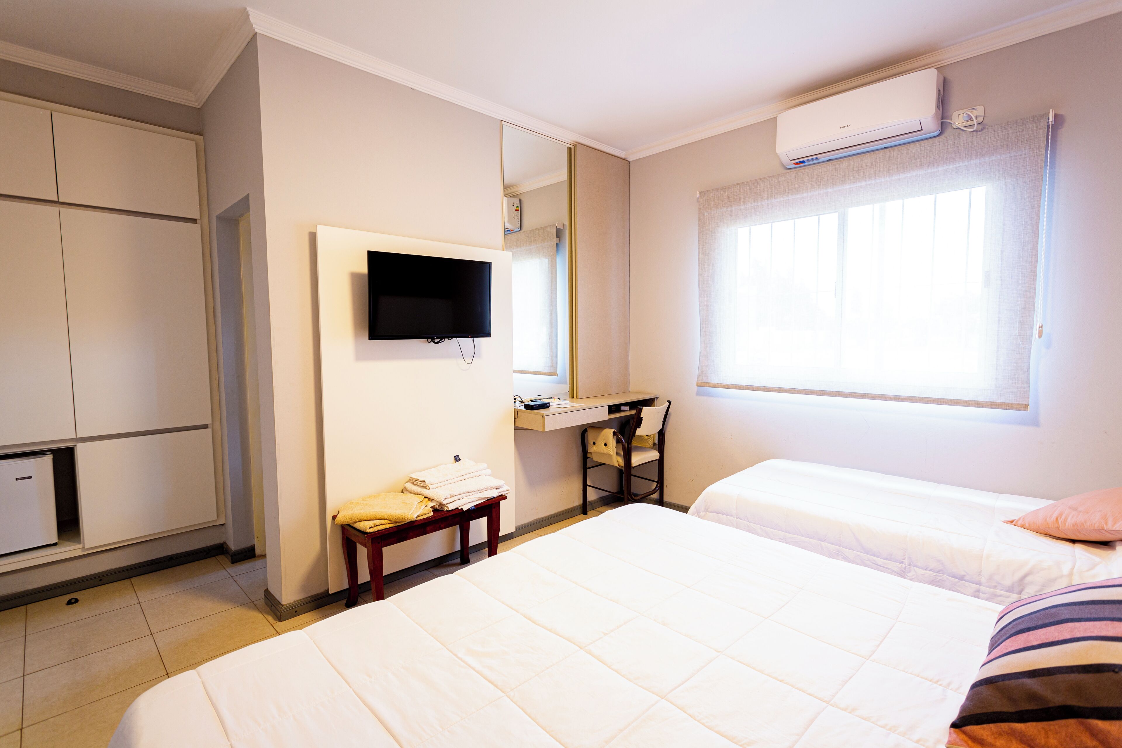 Habitación de hotel con dos camas, televisión, escritorio con silla, espejo y unidad de aire acondicionado.