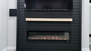 Una sala moderna con un televisor de pantalla plana grande montado en una pared de madera oscura, una chimenea empotrada debajo del televisor y un piso de madera de color claro.