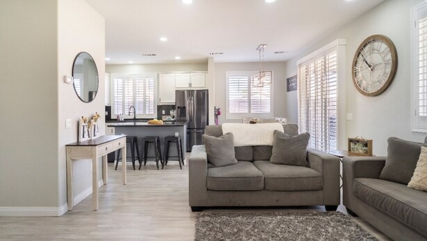 Una sala moderna con un sofá gris, una mesa de centro y una alfombra. Se ve una cocina al fondo con un refrigerador, estufa y fregadero.